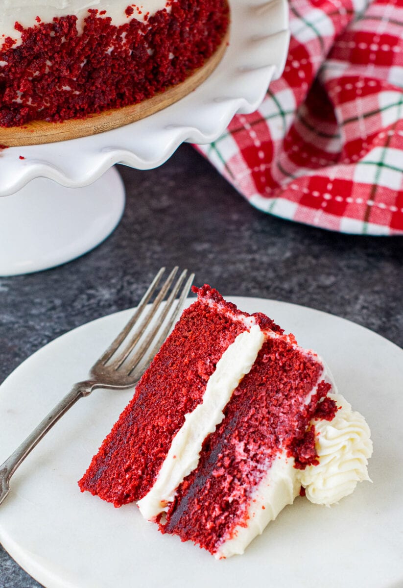Slice of eggless red velvet cake served on a white plate with a cake stand on the side. 